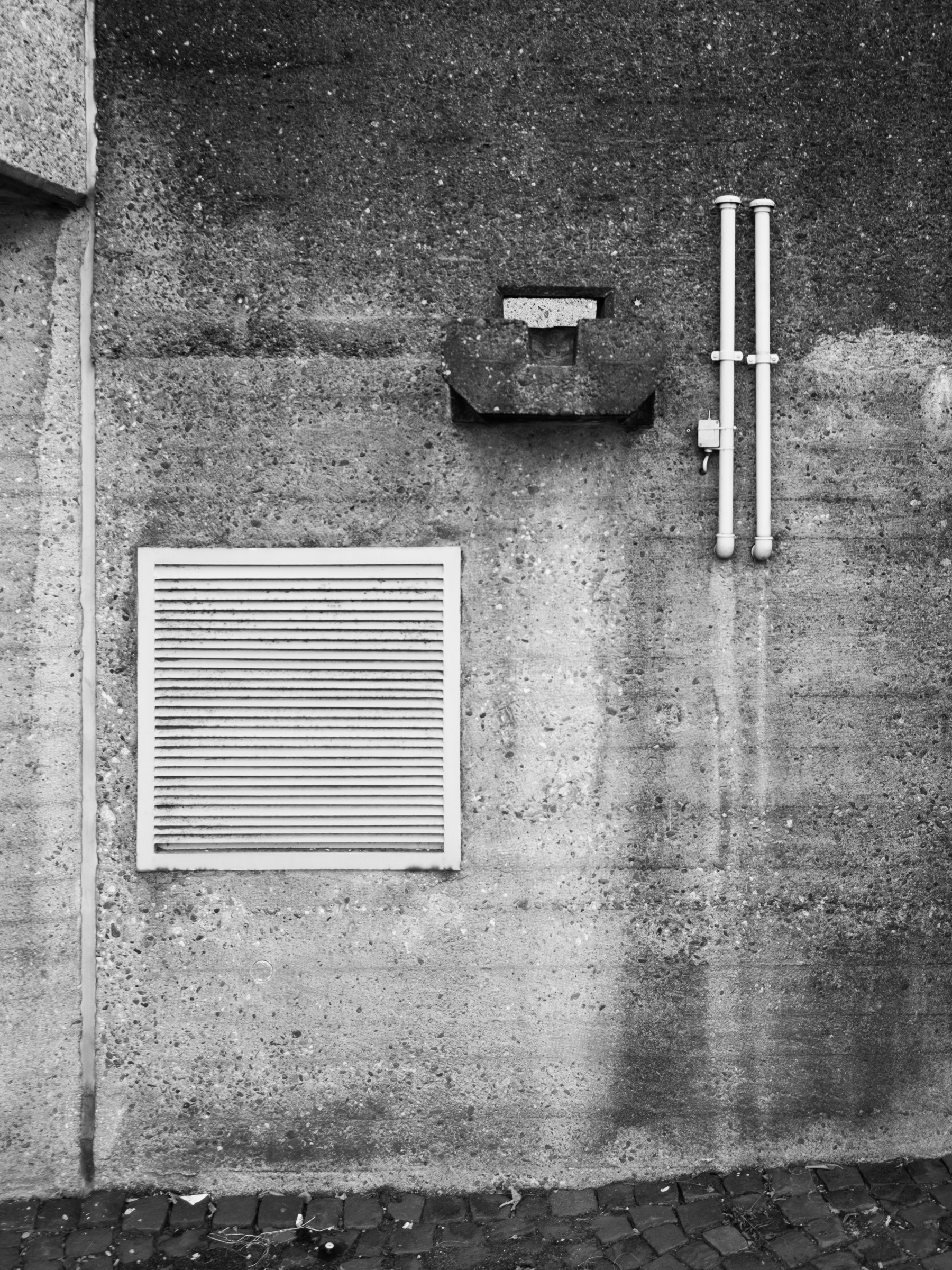 Bare concrete wall detail with ventilation grille, junction box, and pipework