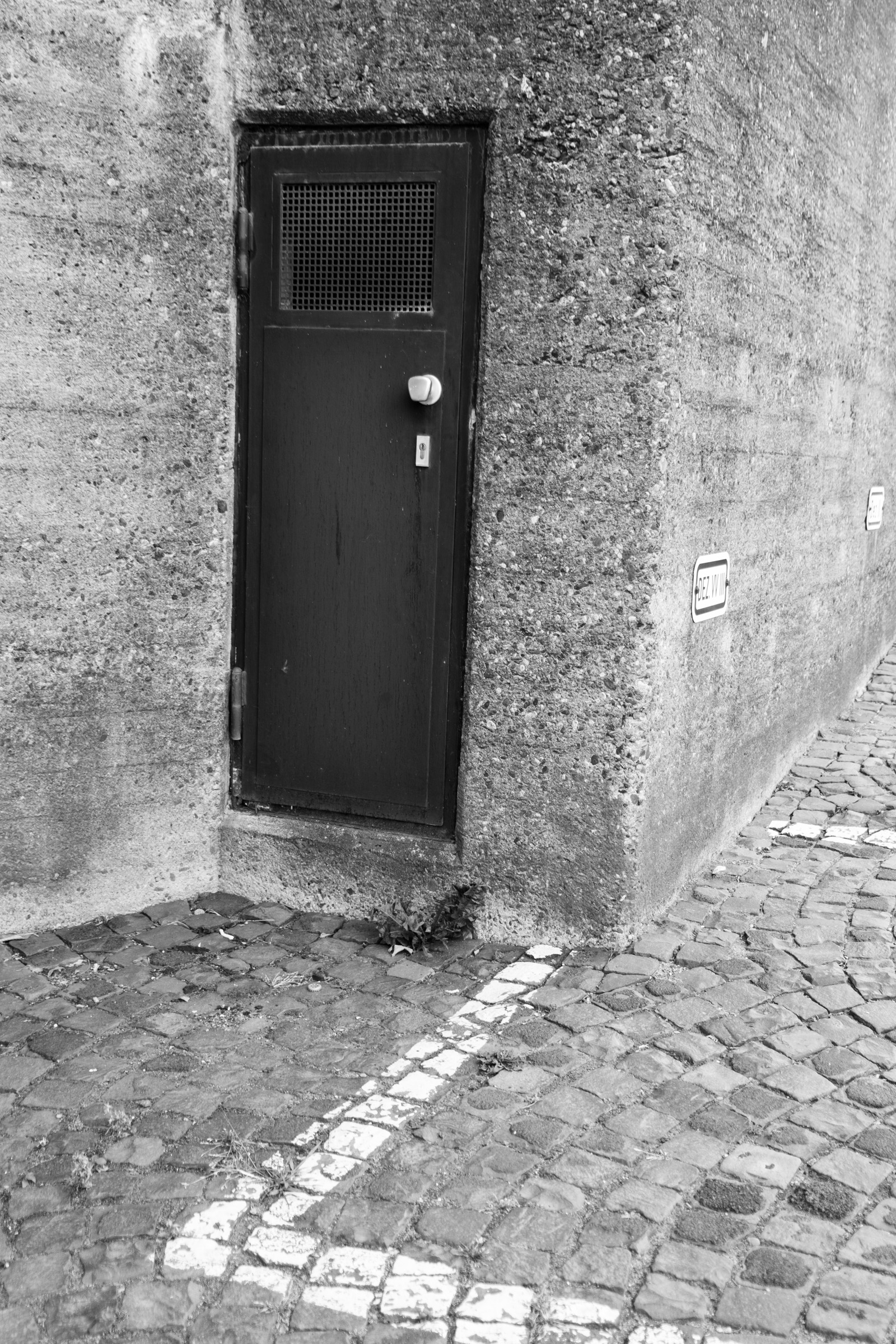 Metal door with mesh grille set into a concrete corner, cobblestone street below