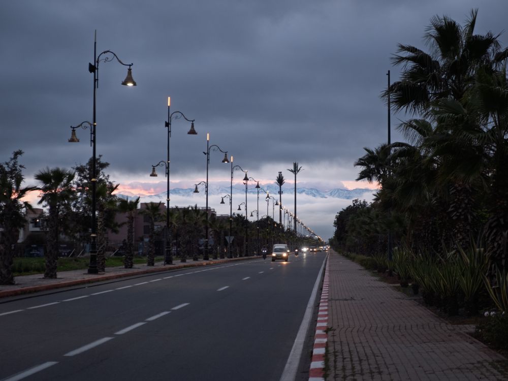 Illuminated street lamps and palm trees on a wide road, with distant mountains under dark clouds.
