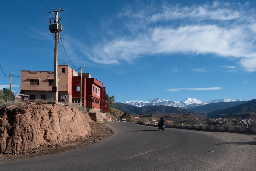 Motorcyclist on a road beside a large, half-finished building, set against snow-capped peaks.