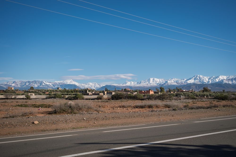 Road leading to a village with snow-capped mountains under a clear blue sky.