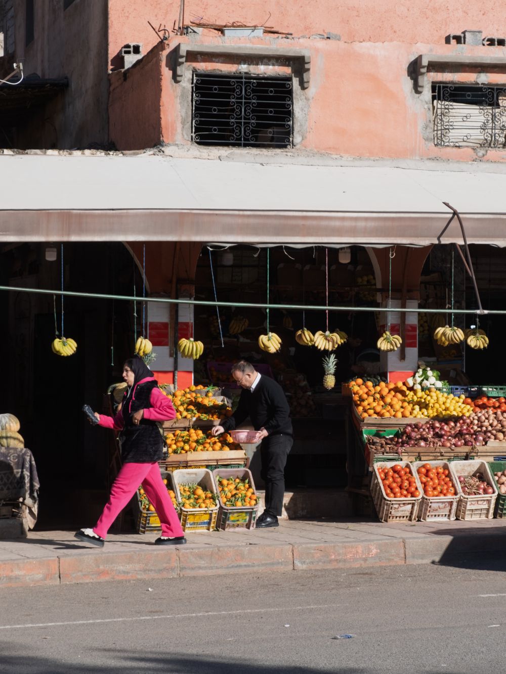 Fruit stand at a street market, with a vendor assisting customers in front of a pink building.